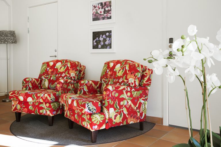 två rödmönstrade fåtöljer står på rund matta, vit orkidé i förgrund två tavlor på väggen