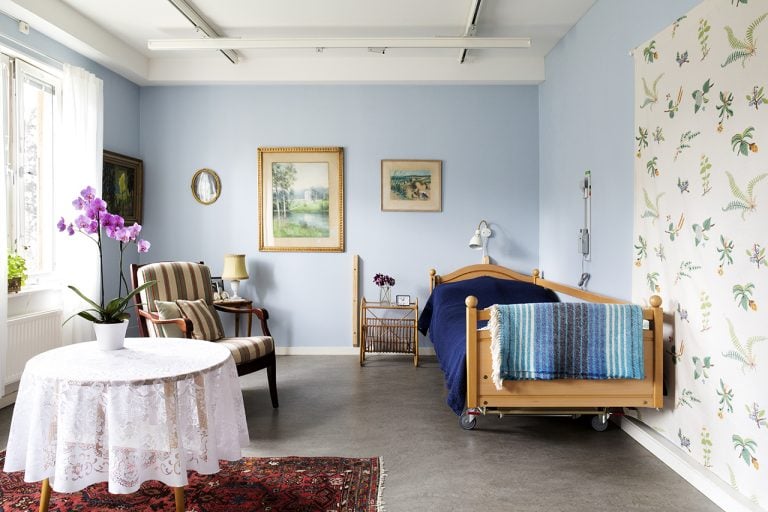 lägenhet delvis mönstrad tapet, säng med blått överkast, runt bord med spetsduk, fåtölj tavlor på vägg