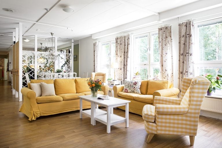gemensamt rum med soffgrupp i gult, litet vitt bord, mönstrade gardiner framför fönster