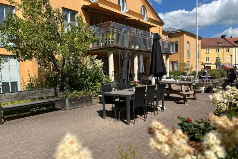 Utemöbler på grusplanen utanför boendets orange fasad. Blommande buskage i förgrunden.