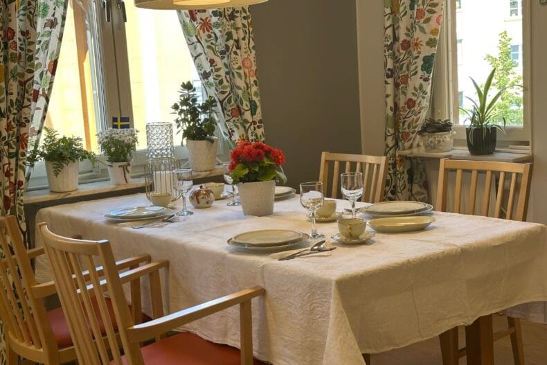 Matbord uppdukat med fint porslin. Hemtrevligt rum med blommiga gardiner och flera krukväxter.