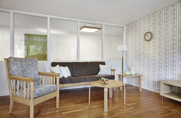 gemensamt rum, svart soffa, två kaffebord, fåtölj, golvlampa i hörnet, glas vägg i bakgrunden som visar korridor