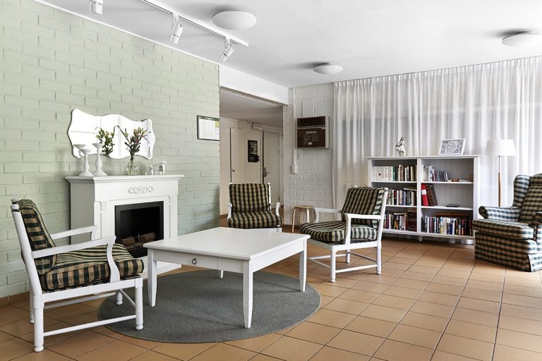 inomhusmiljö, gemensamt vitt rum, ett bord och tre stolar framför en vit eldstad, bokhylla och fotölj i bakgrunden framför ett grå draperi