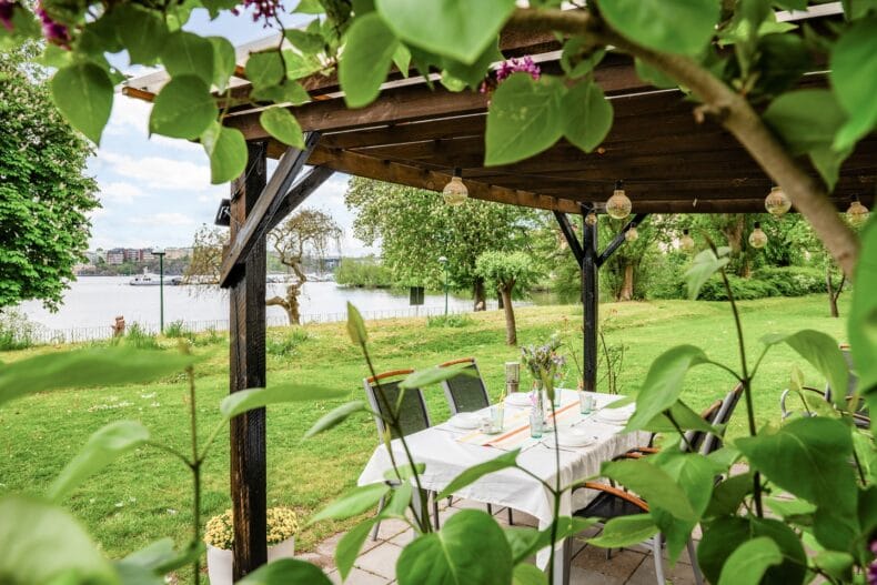 Berså på uteplatsen med festligt dukat bord med vit duk och utelampor i taket med utsikt mot vattnet