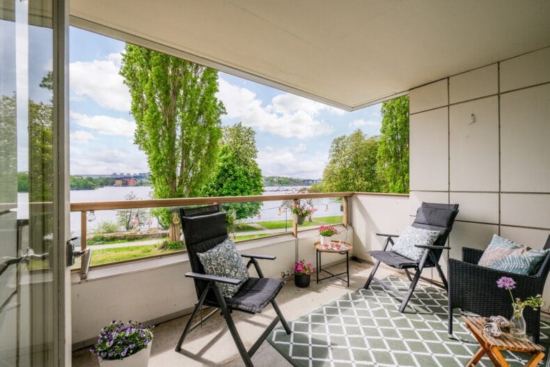 Balkong med tak med rutmönstrad matta och tre svarta utefåtöljer med utsikt över träd och vatten