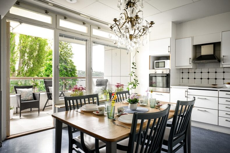 Kök med dukat bord i ek och fyra svarta stolar, kristallkrona i taket och vita köksluckor och bänk. Balkongdörr öppen med utsikt över vattnet
