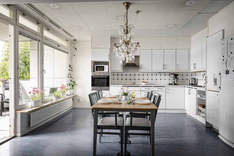 Kök med dukat bord i ek och fyra svarta stolar, kristallkrona i taket och vita köksluckor och bänk.