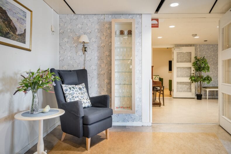 Grå fåtölj med mönstrad kudde, sidobord med blommor, tavla på väggen, vitt vitrinskåp med glas i på väggen med