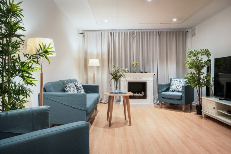 TV-rum med grå soffa och fåtöljer runt bord i ek två golvlampor växter samt vit eldstad