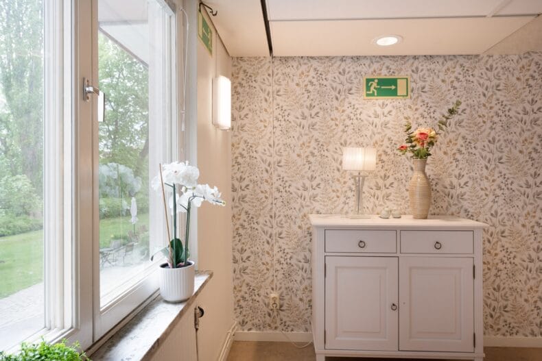 Fönster med orkidé på fönsterbrädet vit byrå med lampa och vas ovanpå mönstrad tapet/fondvägg