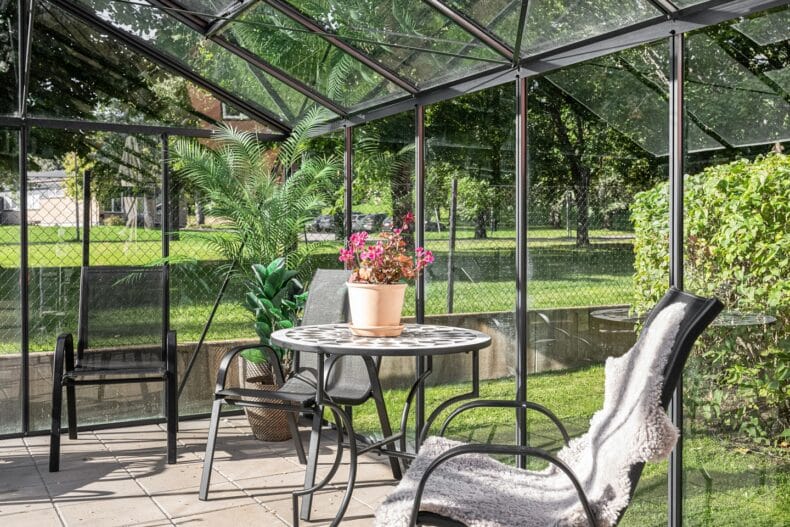 Stol med pläd i ett växthus med stenlagt golv och gräsytor utanför.