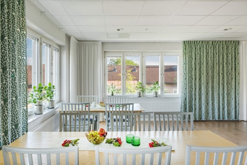 Matsal med flera matbord. Fönster i två väderstreck med gröna mönstrade gardiner.