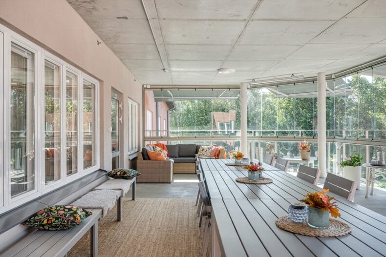 Stor inglasad balkong med långbord i förgrunden och soffhörna i bakgrunden. Utsikt mot grönskande träd.