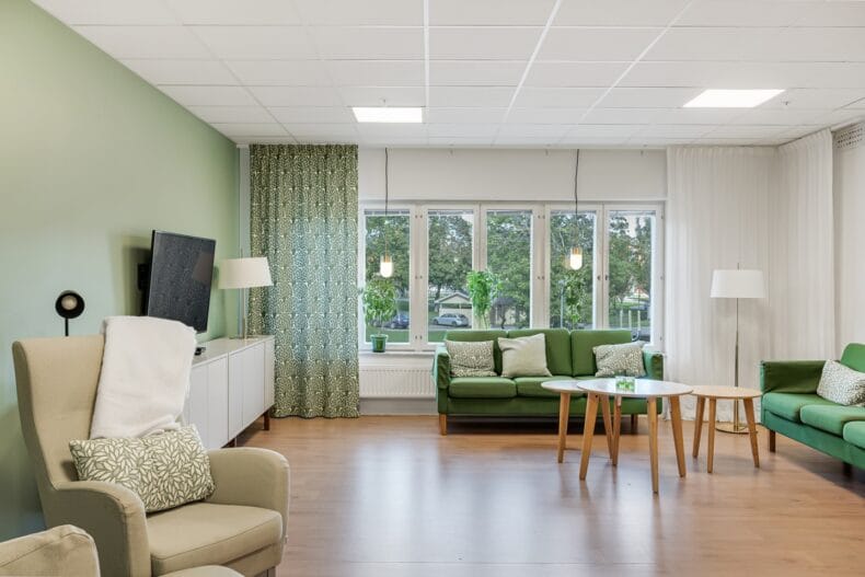 TV-hörna i rum med gröna soffor och fåtöljer samt grön vägg. Fönster mot gräsmatta och stora lövträd.