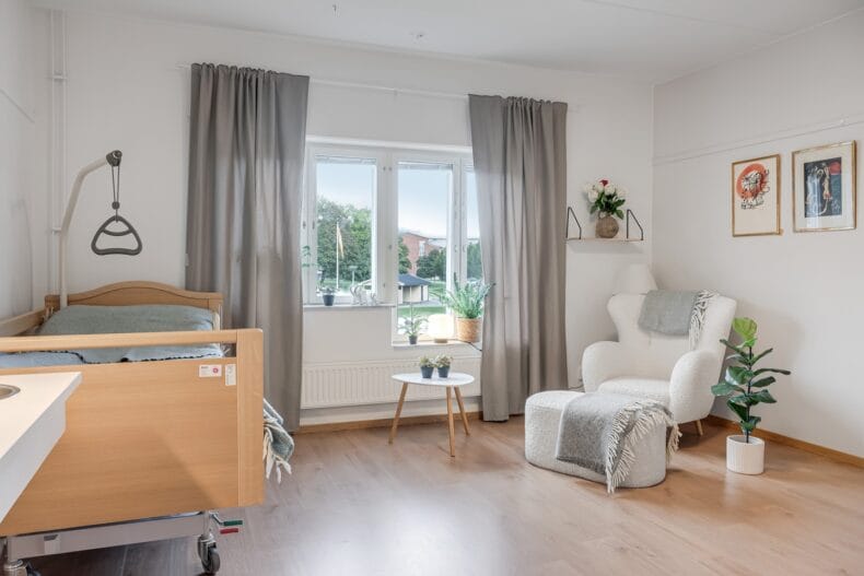 Lägenhet med justerbar säng till vänster och bekväm fåtölj med fotpall till höger. I mitten ett fönster med grå gardiner.