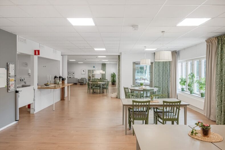 Luftig matsal med flera bordsgrupper. Till vänster en disk in till köket, med kaffetermosar uppställda.