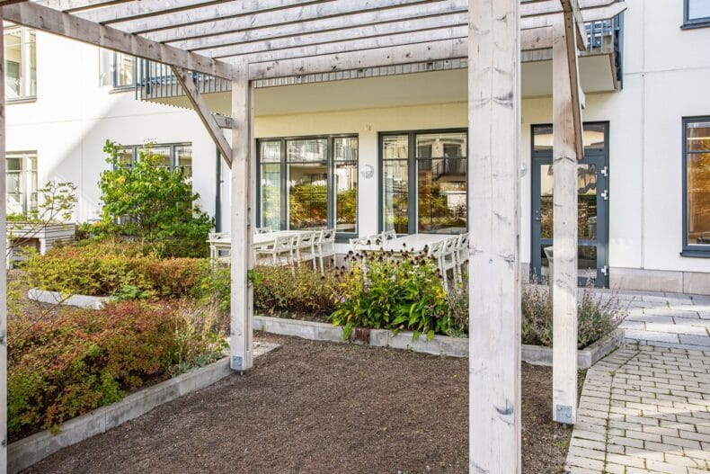 Pergola på innergården och två långbord under tak bakom rabatter med blommor.