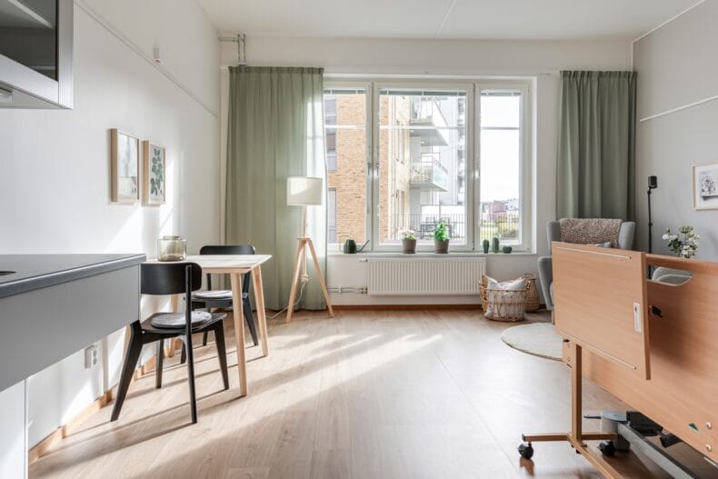 Lägenhet med justerbar säng i förgrunden, matplats till vänster och fåtölj till höger i bakgrunden. Stora ljusa fönster med gröna gardiner.