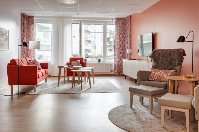 TV-hörna i ett rum med röda textilier och röd vägg. Soffa och fåtölj framför stora ljusa fönster.