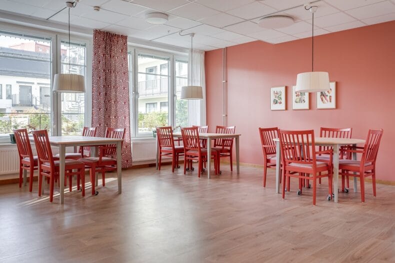 Matsal med flera bordsgrupper. Stolarna har hjul på främre benen. Till vänster höga ljusa fönster med växter utanför.