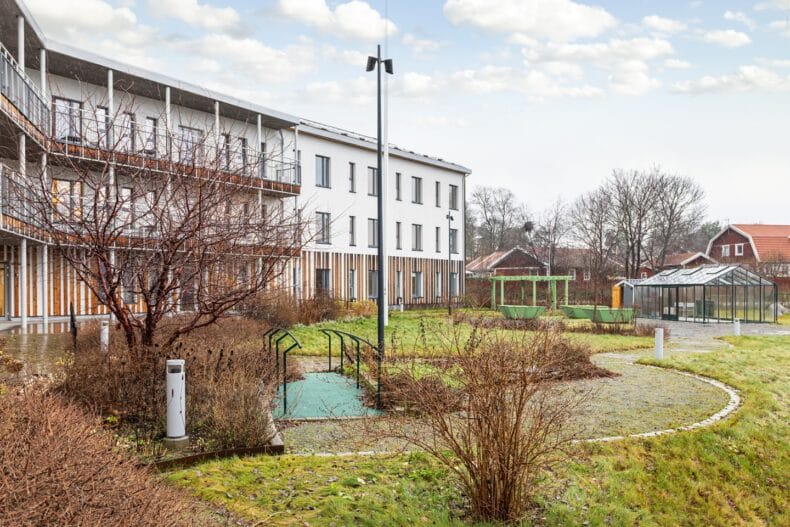 En modern flerbostadsbyggnad med tre våningar omgiven av grönska, gångvägar och planteringar i en lugn gårdsmiljö.
