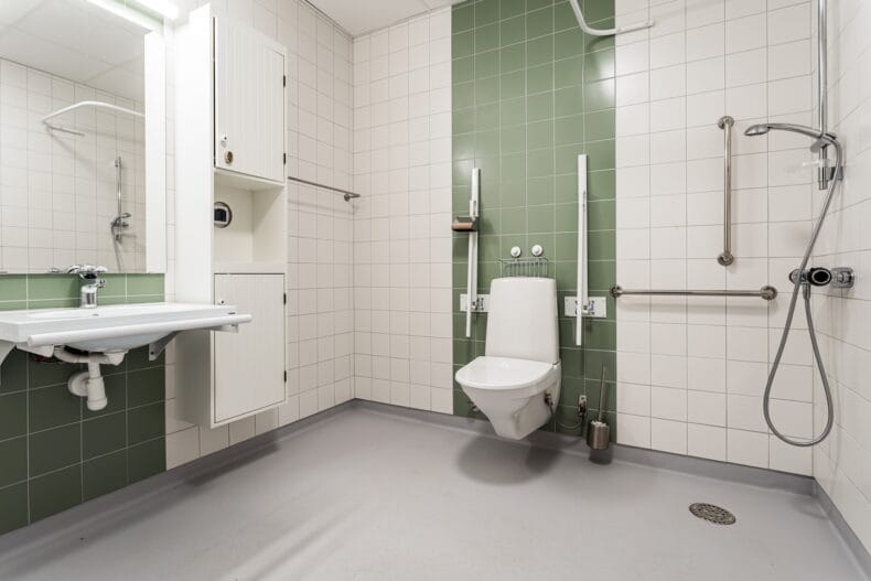 Ett tillgänglighetsanpassat badrum med toalett, handfat, dusch och stödhandtag mot kaklade väggar i vitt och grönt.
