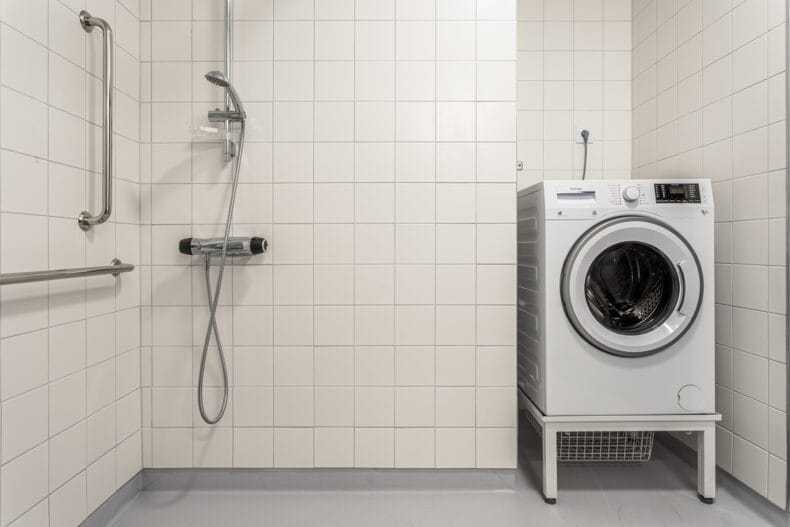 En duschplats i ett kaklat badrum med väggmonterad dusch och en tvättmaskin placerad intill.