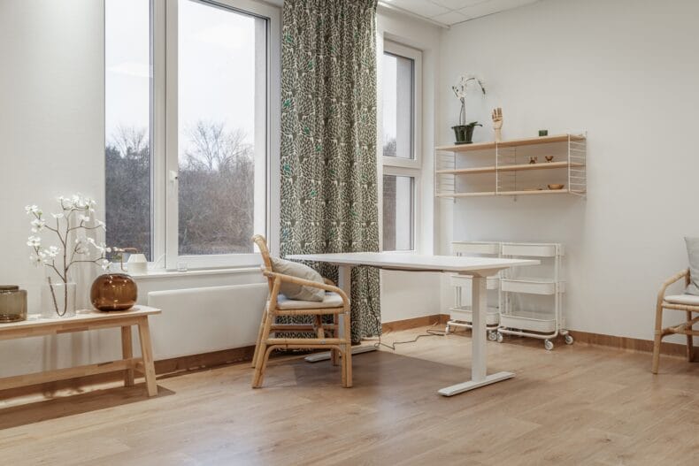 Ett ljust rum med stora fönster, bord, stolar och förvaring på hjul, inrett med växter och textilier.