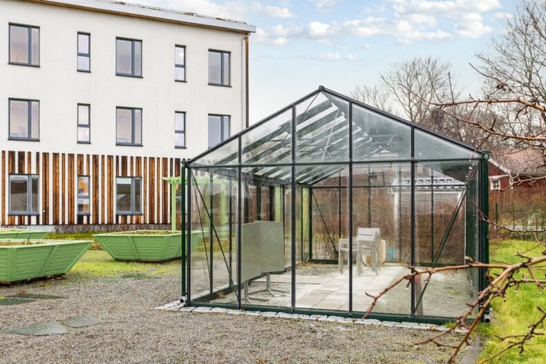 Ett växthus i glas placerat på en grusad yta intill gröna odlingslådor, med en ljus byggnad bakom.