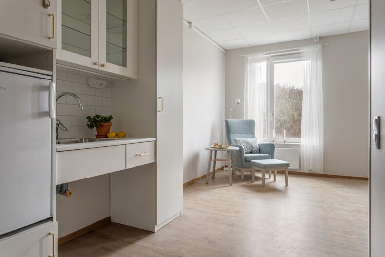 Ett rum där ett pentry med diskho och kylskåp syns i förgrunden och en fåtölj med fotpall står vid ett fönster.