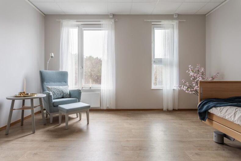 Ett ljust rum med en fåtölj och fotpall vid fönstret samt en säng placerad längs väggen.