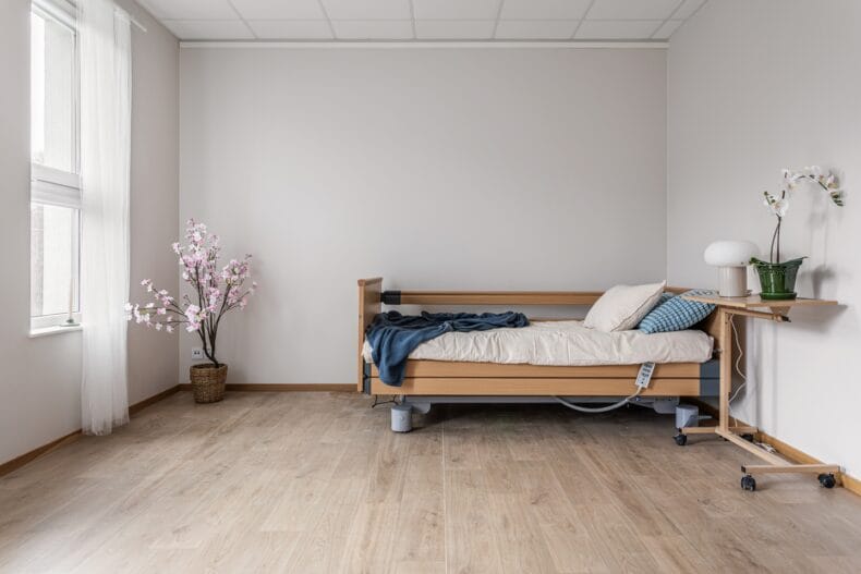 En justerbar säng i ett ljust rum med trädetaljer, nattduksbord och en blommande växt i kruka.