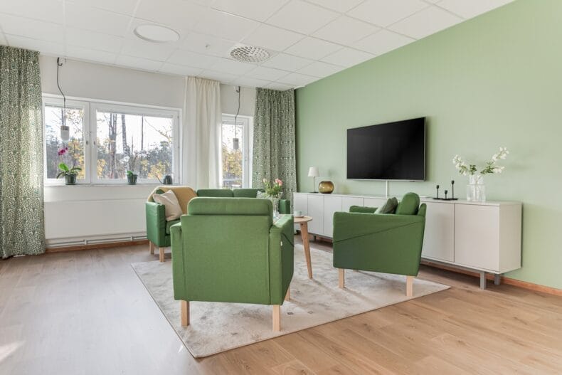 Ljust vardagsrum med gröna fåtöljer, tv på väggen och stora fönster som släpper in dagsljus.