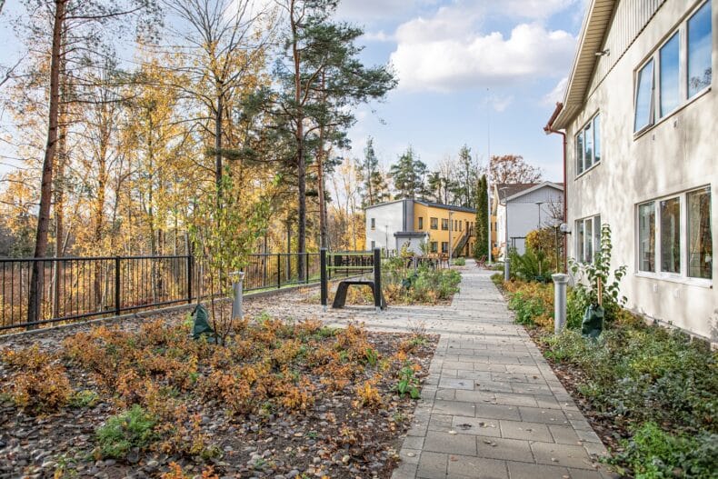 Gångväg genom trädgård med höstlöv, planteringar och bostadshus i bakgrunden.