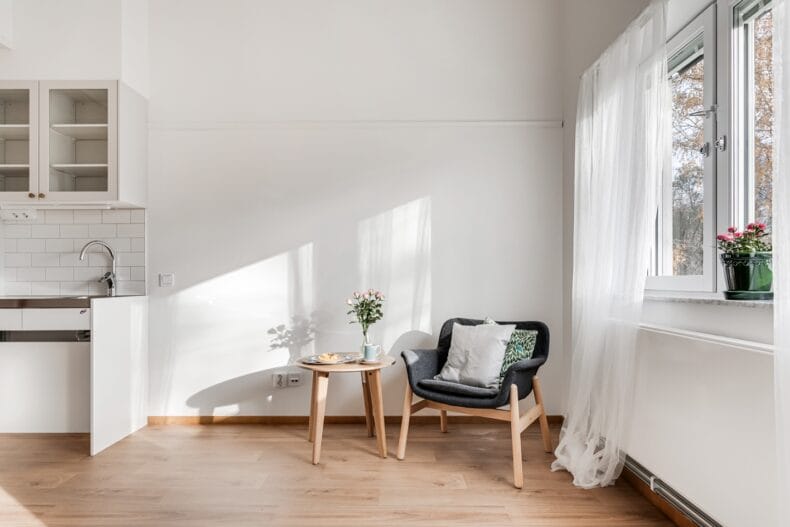 Närbild av sittgrupp med fåtölj, sidobord och ljusinsläpp från fönster. Trinettkök skymtar.