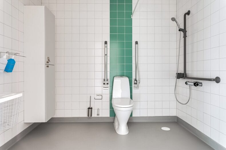 Tillgänglighetsanpassat badrum med vitt kakel och grön accentvägg.