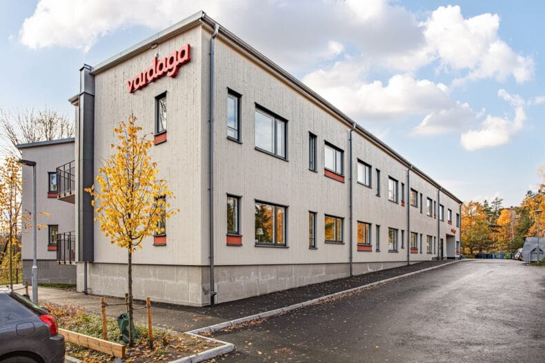 Fasadvy av byggnad med Vardaga-logotyp, stora fönster och träd med höstfärger.