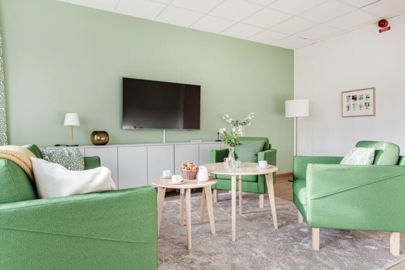 Vardagsrum med gröna fåtöljer och soffor, två runda bord med kaffekoppar och blommor.