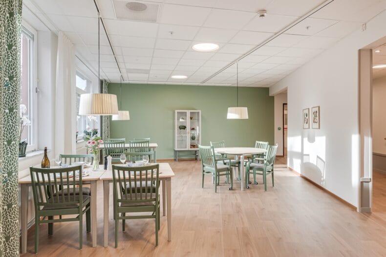 Ljus matsal med gröna stolar, träbord och vita taklampor, med grön vägg i bakgrunden.