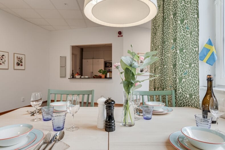 Närbild på dukat matbord med porslin, glas och blommor i vas, med köksö i bakgrunden.