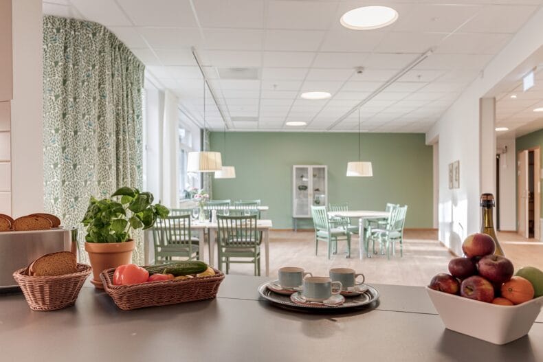 Vy från köket mot matsalen med grön inredning, träbord och stora fönster.