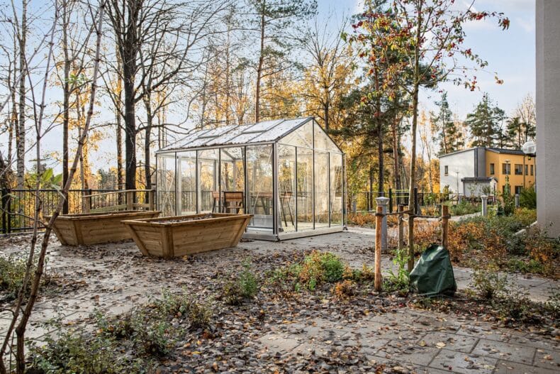 Trädgård med växthus och odlingslådor omgivna av träd i höstfärger.