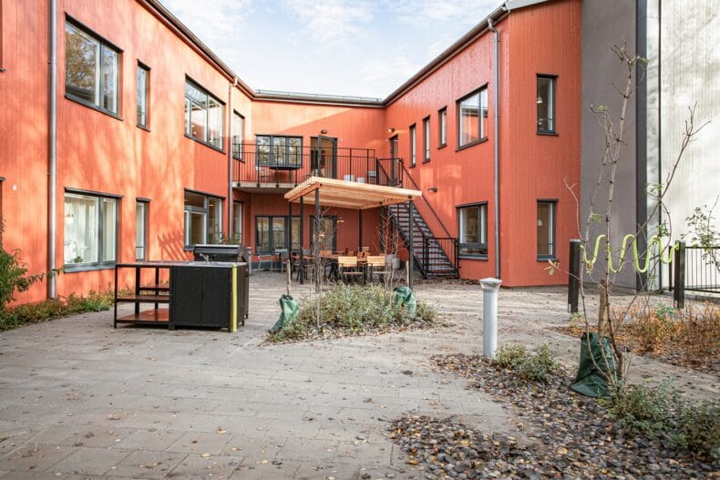 Innergård med röd fasad, utemöbler under pergola och trappor till övervåning.