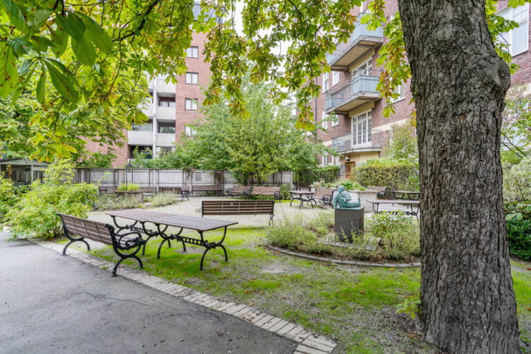 utomhus, parkbänk och parkbord, träd i förgrunden, fontän i mitten, träd och lägenheter i bakgrunden