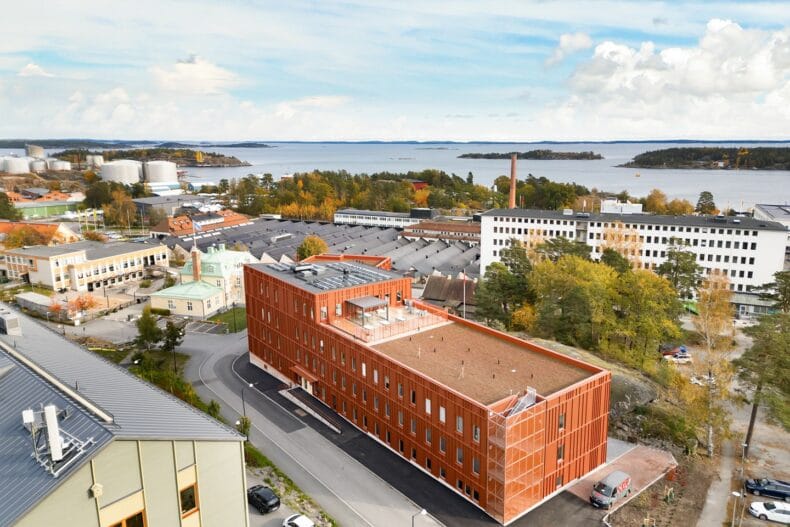 Flygbild över byggnaden och området. En modern tegelröd byggnad omgiven av andra hus och grönska, med utsikt över hav och öar i bakgrunden en solig höstdag.