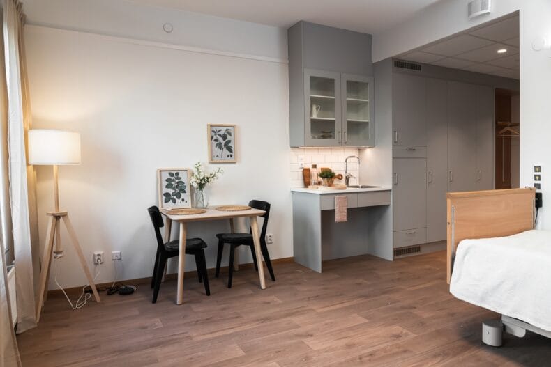 Litet kök och matplats. Ljus och modern lägenhetsinteriör med pentry, matbord med två stolar, tavlor och blommor, samt säng i förgrunden.