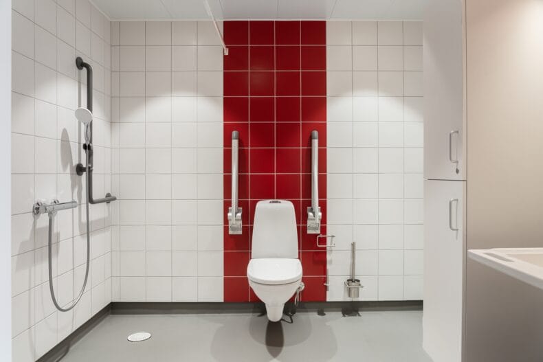 Tillgänglighetsanpassat badrum med vitt kakel, röd accentvägg och stödhandtag vid toaletten.