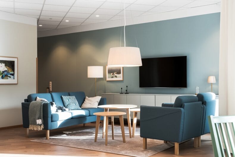 Vardagsrum i blått tema. Ljust vardagsrum med blå soffor och fåtöljer, ljusa träbord och dämpad röd vägg bakom tv:n.