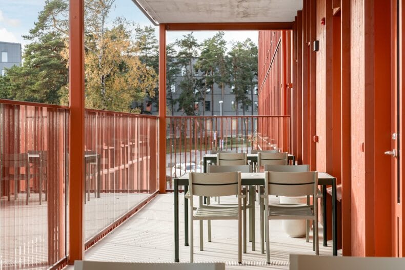 Uteplats med sittplatser. Täckt balkong med röda väggar och flera bord med stolar, omgiven av träd och grönska.