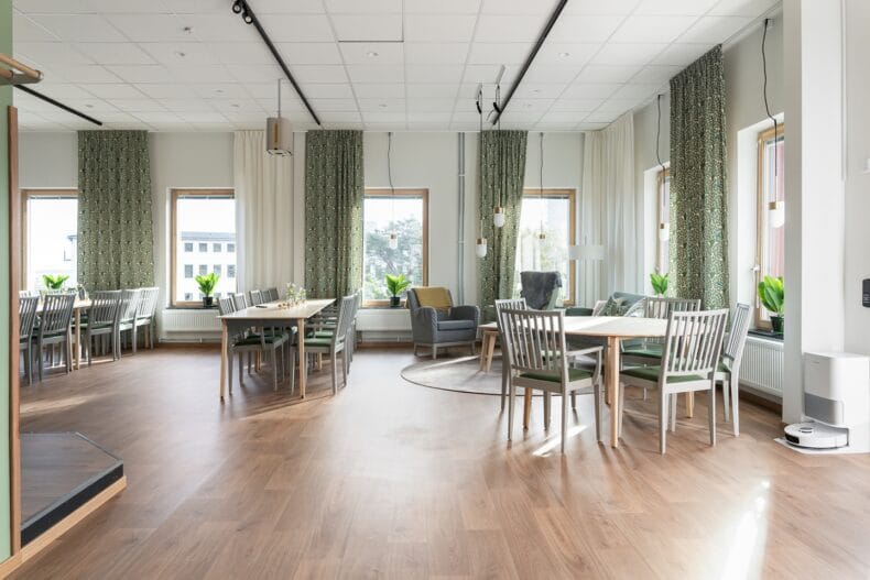 Matsal med stora fönster. Ljus matsal med bord och stolar, stora fönster med grönmönstrade gardiner och växter i fönstren.